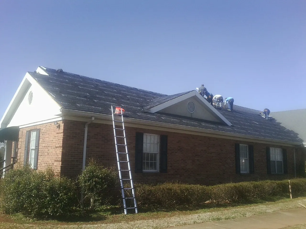 Crew tearing shingles off a brick ranch roof with a ladder set — Emergency Roof Repair work by our crew in West Concord
