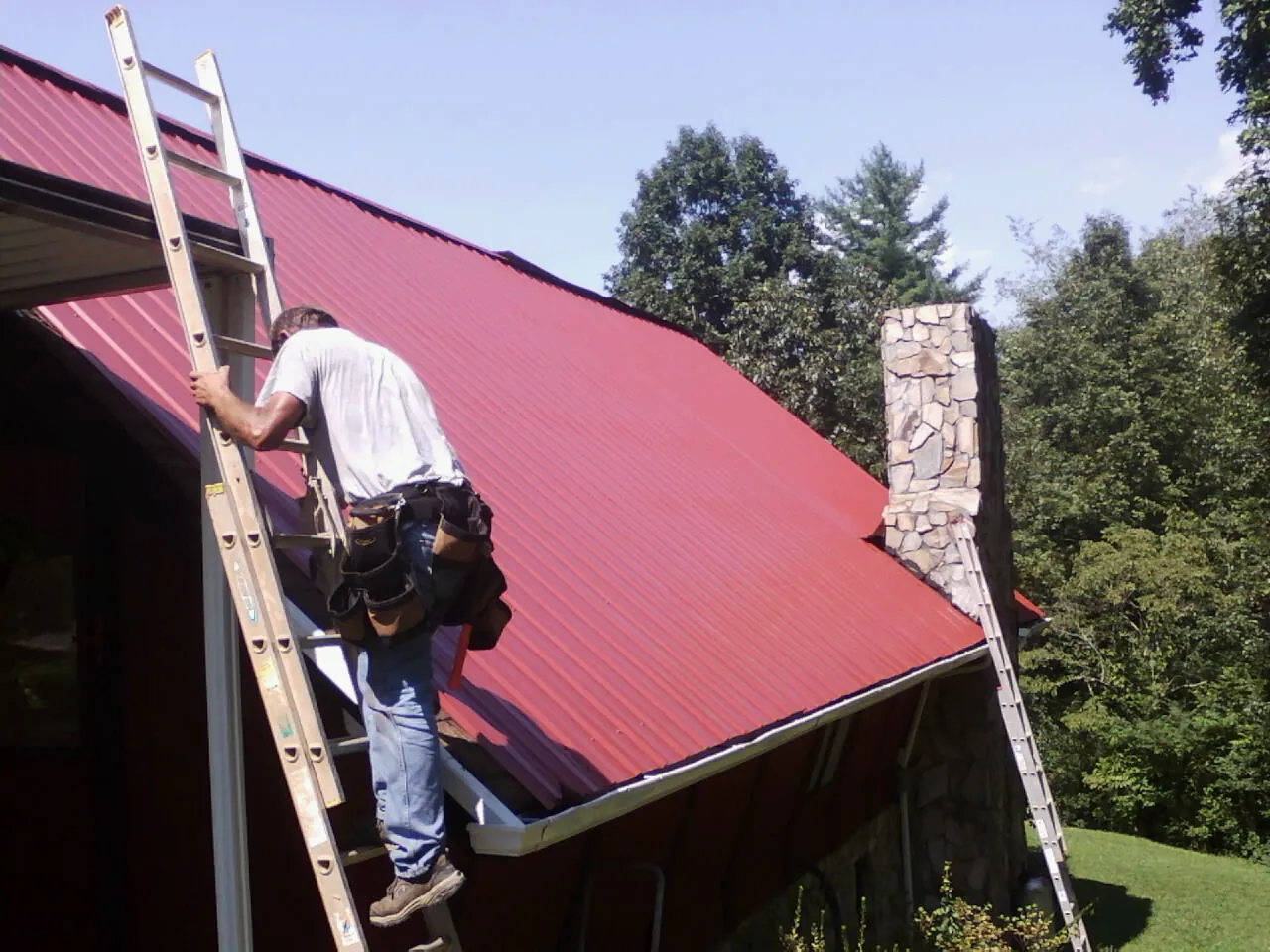 Roofer on a ladder beside a red metal roof and stone chimney — Soffit & Fascia Repair by Roofing Brothers in West Concord